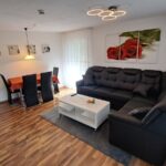 Modernes Wohnzimmer der Ferienwohnung Ahorn mit schwarzer Eckcouch, Esstisch und großen Fenstern, die viel Tageslicht hereinlassen. Ein idealer Raum zum Entspannen.