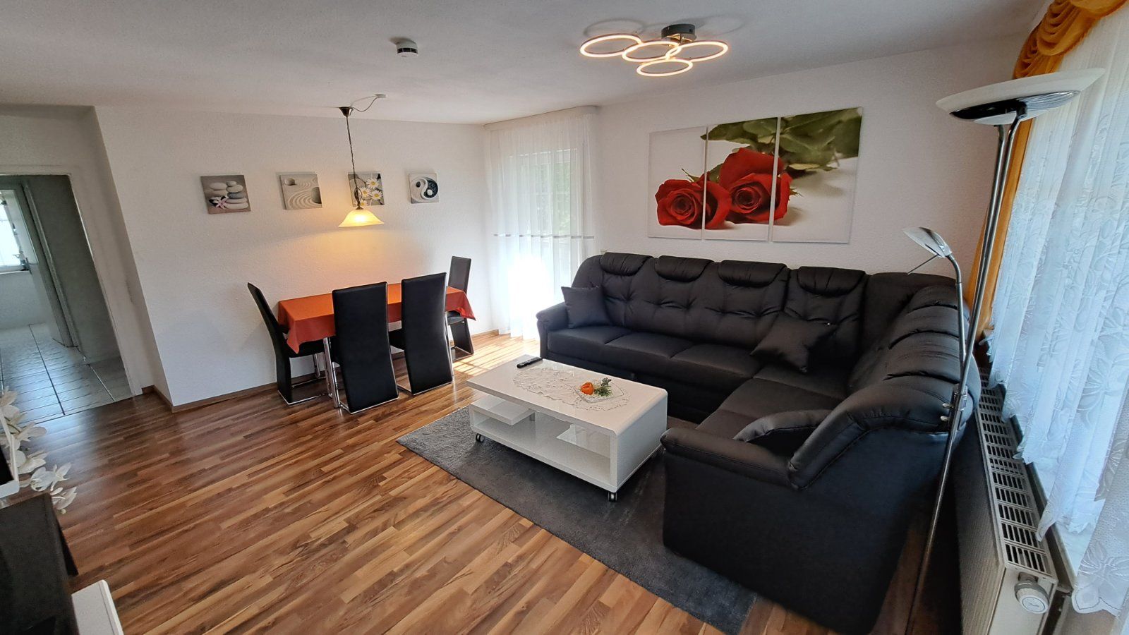 Modernes Wohnzimmer der Ferienwohnung Ahorn mit schwarzer Eckcouch, Esstisch und großen Fenstern, die viel Tageslicht hereinlassen. Ein idealer Raum zum Entspannen.