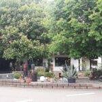 Außenansicht eines Restaurants oder Geschäfts in Küssaberg, umgeben von Bäumen. Der Bereich hat eine Terrasse mit Tischen und Stühlen und ein Schild zeigt den Standort an.