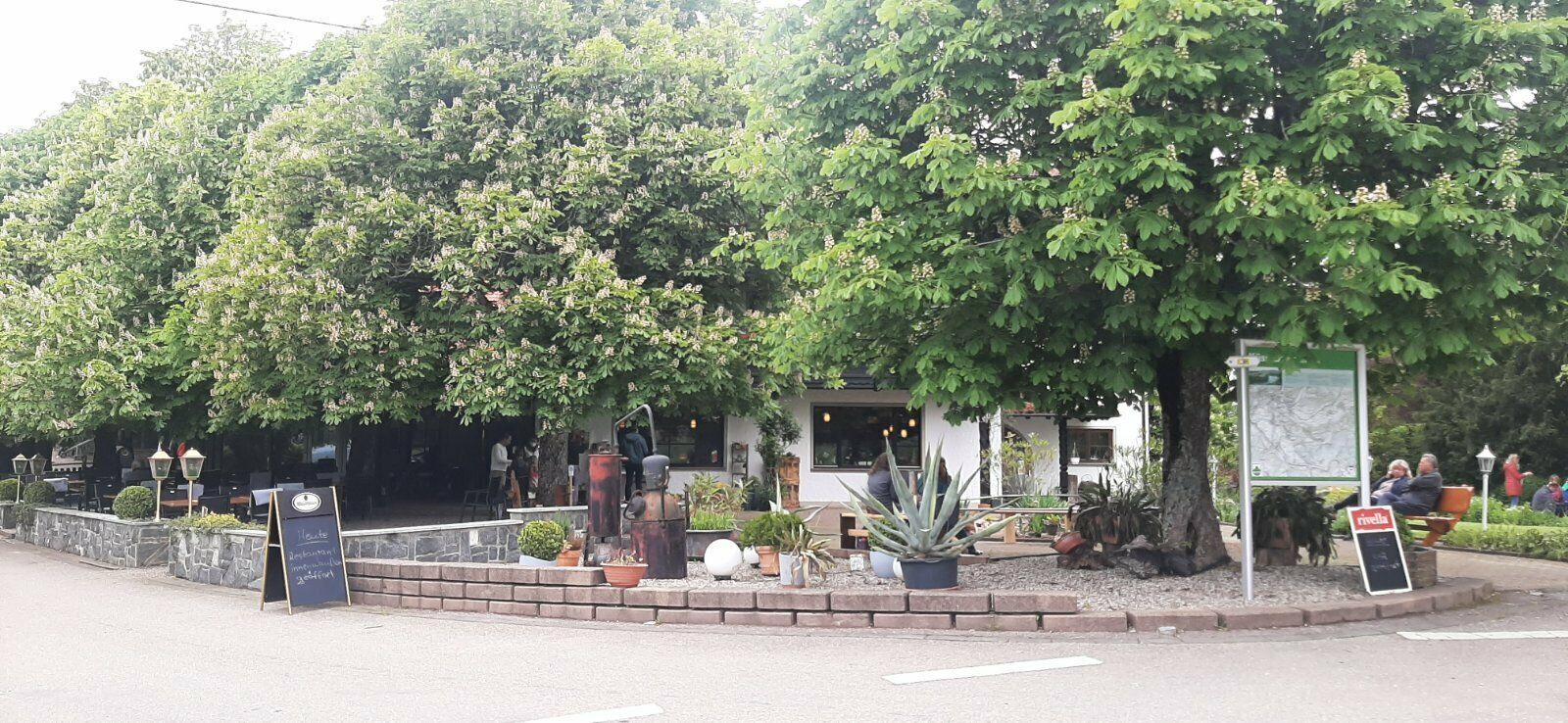 Außenansicht eines Restaurants oder Geschäfts in Küssaberg, umgeben von Bäumen. Der Bereich hat eine Terrasse mit Tischen und Stühlen und ein Schild zeigt den Standort an.