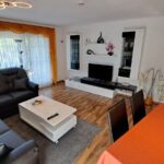 Gemütliches Wohnzimmer der Ferienwohnung Ahorn mit schwarzer Eckcouch, Esstisch und modernen Möbeln. Große Fenster lassen viel Tageslicht herein und schaffen eine angenehme Atmosphäre.