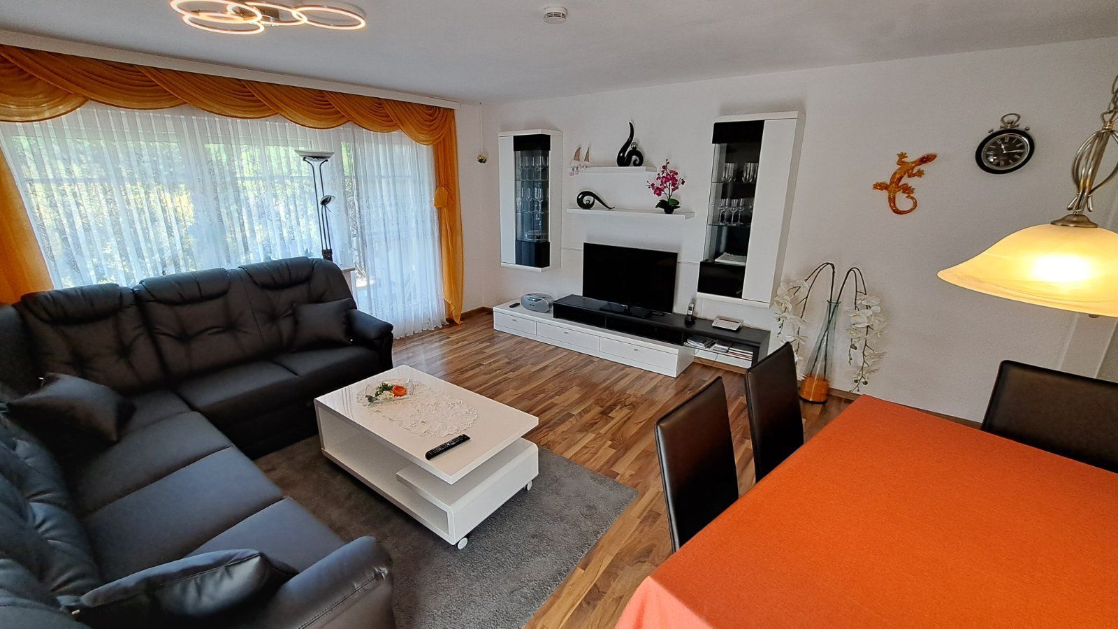Gemütliches Wohnzimmer der Ferienwohnung Ahorn mit schwarzer Eckcouch, Esstisch und modernen Möbeln. Große Fenster lassen viel Tageslicht herein und schaffen eine angenehme Atmosphäre.