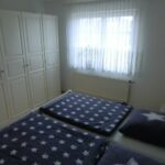 Schlafzimmer in der Ferienwohnung Ahorn mit zwei Einzelbetten und einem großen Kleiderschrank. Das Bettzeug ist mit einem Sternenmuster versehen, und das Zimmer ist mit viel Tageslicht durch ein großes Fenster beleuchtet.