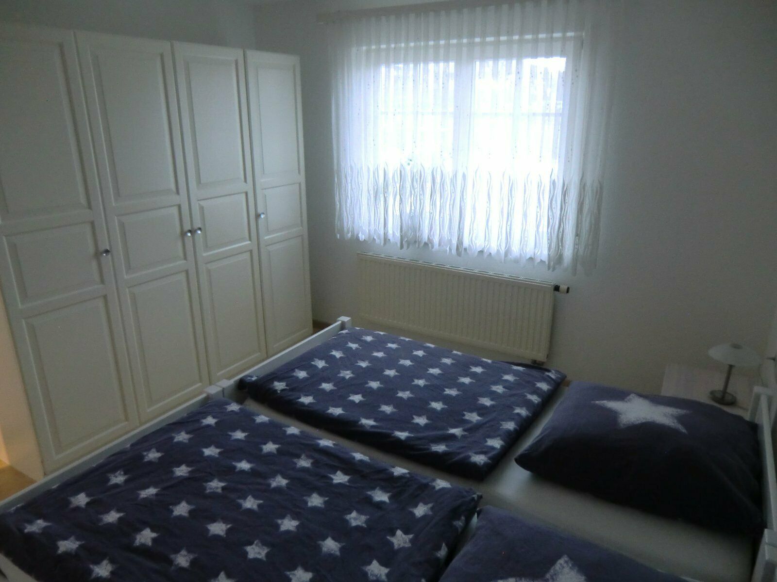 Schlafzimmer in der Ferienwohnung Ahorn mit zwei Einzelbetten und einem großen Kleiderschrank. Das Bettzeug ist mit einem Sternenmuster versehen, und das Zimmer ist mit viel Tageslicht durch ein großes Fenster beleuchtet.